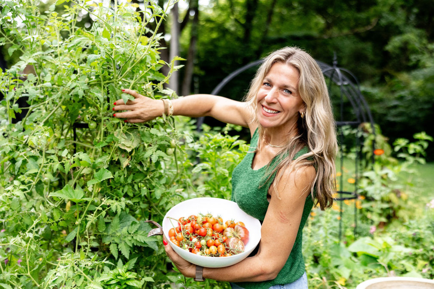 Smiling woman harvesting cherry tomatoes from a lush garden, holding a white bowl filled with freshly picked tomatoes in a vibrant backyard setting