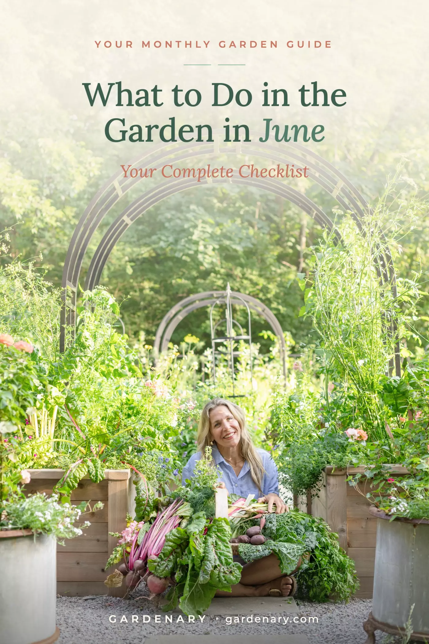 Nicole Johnsey Burke sitting in her kitchen garden surrounded by cedar raised beds, holding a harvest basket of beets, kale, and vegetables, with the Gardenary Nicole Arch Trellis in the background — What to Do in the Garden in June checklist