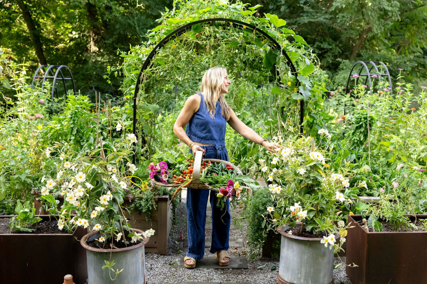 Woman holding a harvest basket filled with fresh vegetables and flowers, standing under a garden arch surrounded by lush raised garden beds.