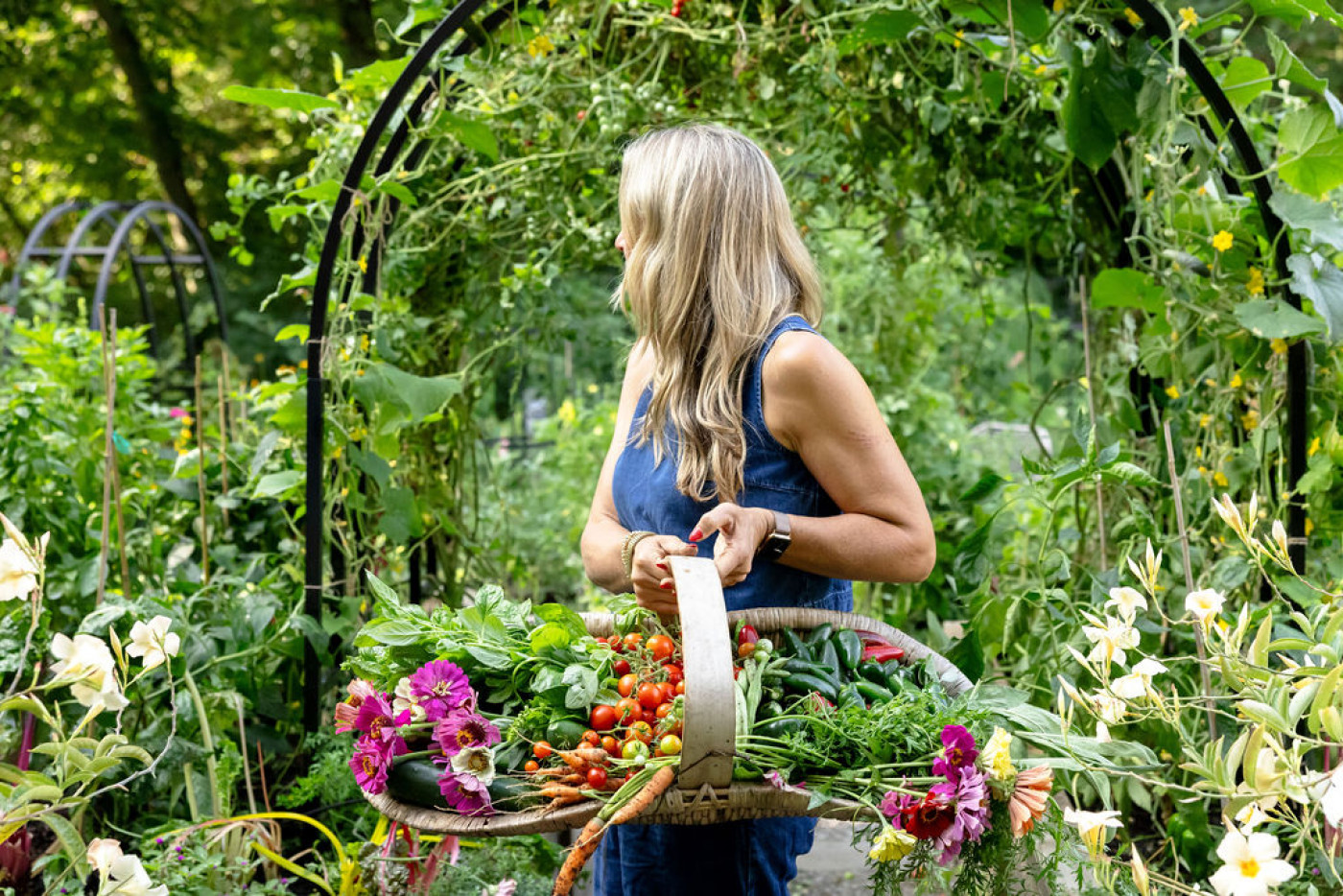 gardener with harvest basket full of zinnias, cucumbers, carrots, and tomatoes standing in garden